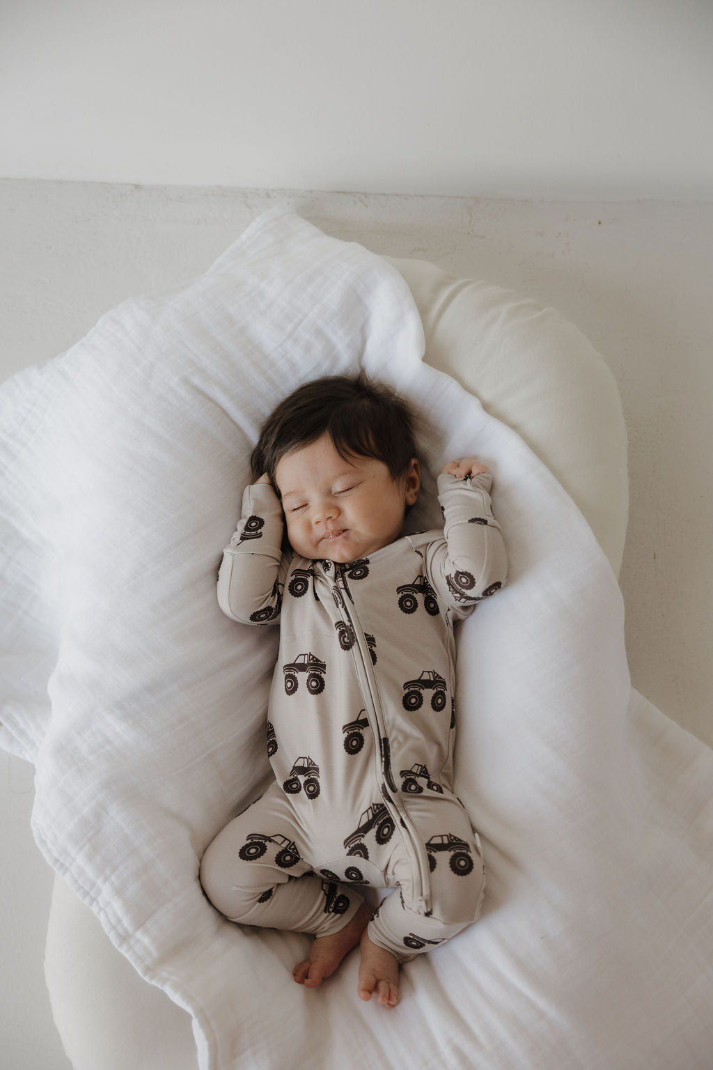 A dark-haired baby sleeps peacefully on a white blanket, wearing forever french baby Bamboo Zip Pajamas in the Monster Truck print, arms raised near their head.