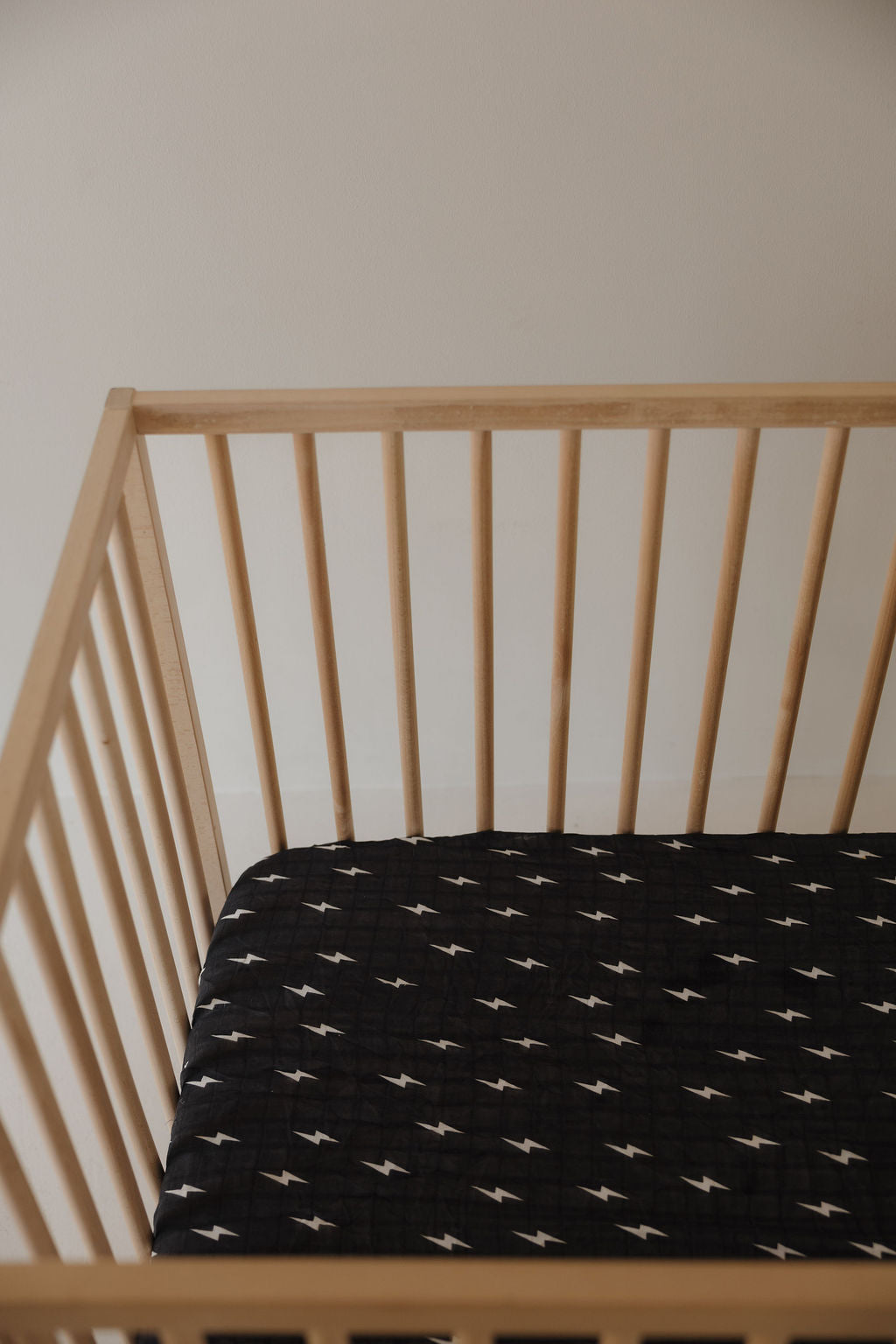 The forever french baby Muslin Crib Sheet | Midnight Bolt features a stylish white lightning bolt pattern on black, shown on a wooden crib against a light wall for chic and comfortable nursery bedding.