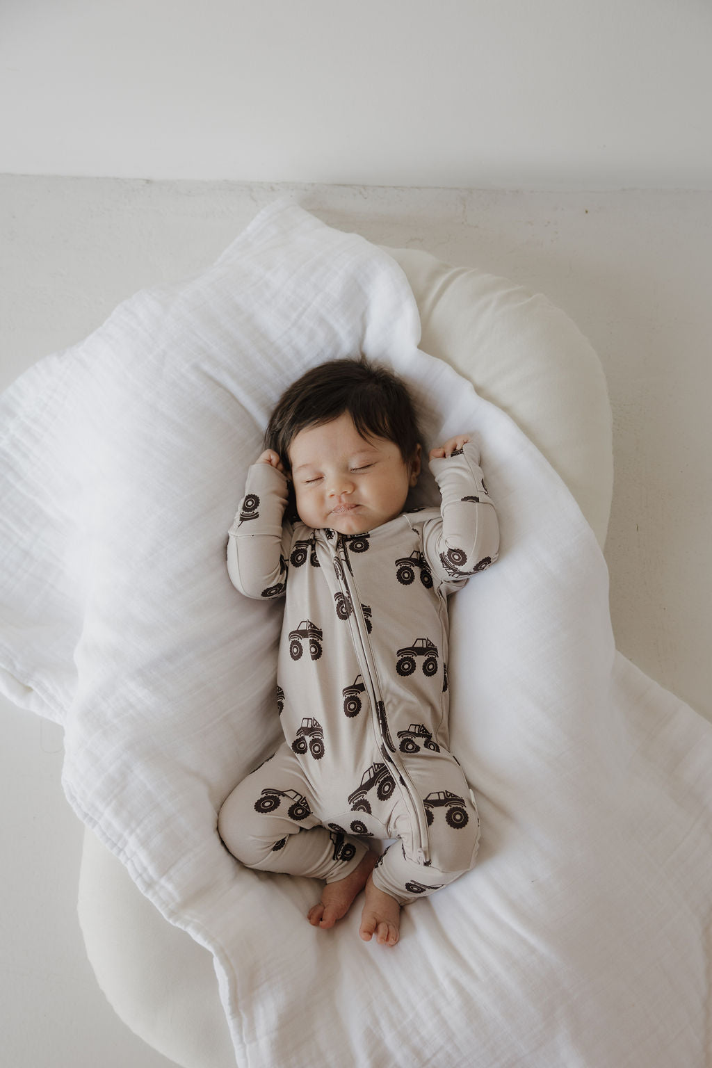 A baby with dark hair sleeps peacefully on a white surface, wrapped in a soft blanket and wearing forever french baby's Bamboo Zip Pajamas | Monster Truck, a beige, breathable onesie featuring black monster trucks.