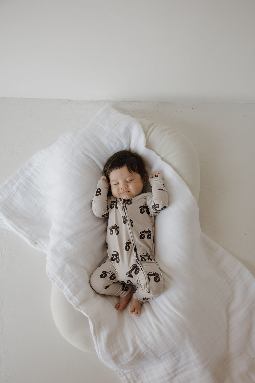 A baby with dark hair sleeps peacefully on a white blanket, wearing forever french baby Bamboo Zip Pajamas in the Monster Truck print. The light, breathable fabric features playful monster truck designs and keeps the baby's bare feet comfy.