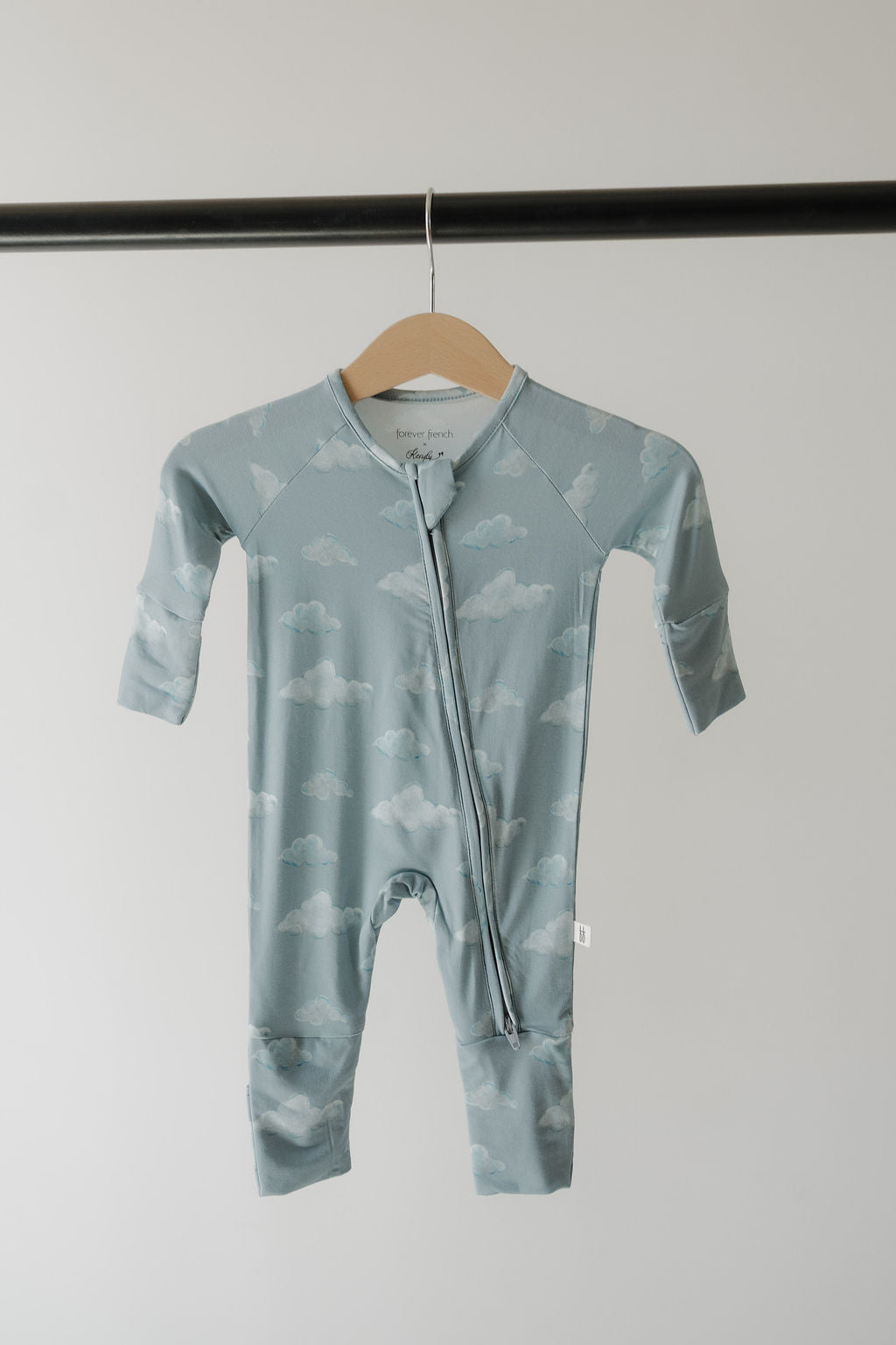 The Kendy 3 Bamboo Zip Pajamas | Duke features a light blue, cloud-patterned design with long sleeves and legs. Made from hypo-allergenic, breathable fabric, it has a diagonal zipper—perfect for sensitive skin. Shown on a wooden hanger.