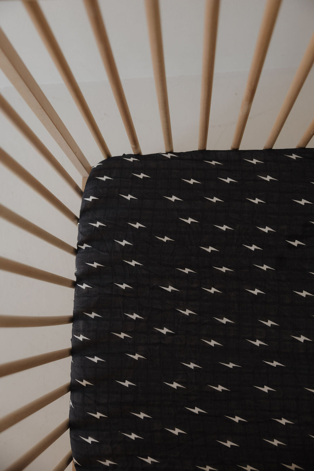 A wooden crib with vertical slats and a forever french baby Muslin Crib Sheet | Midnight Bolt—breathable black muslin with a white lightning bolt pattern—for stylish baby bedding.