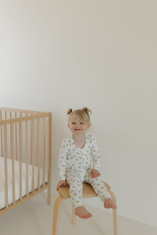 A young child with blonde pigtails sits on a wooden chair, wearing the Bamboo Two Piece Pajamas in Daisy Fields by forever french baby. The minimalistic room has light walls, with a wooden crib and white mattress in the background.