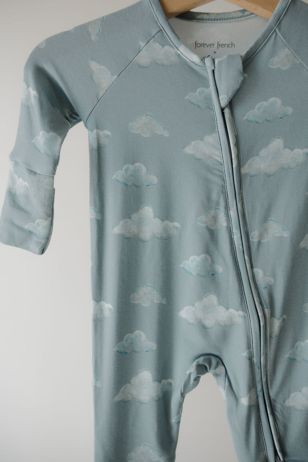 The Kendy 3 Bamboo Zip Pajamas | Duke, a light blue baby onesie with white cloud patterns, features breathable fabric, a front zipper, and a fold-over collar. Displayed on a wooden hanger against a plain background.
