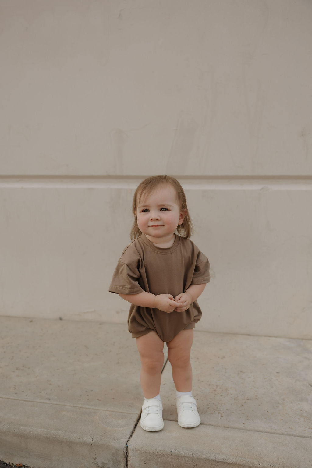 A toddler with light brown hair smiles at the camera while wearing the forever french baby Short Sleeve Romper in Patina and white shoes, standing on a concrete surface in front of a beige wall.