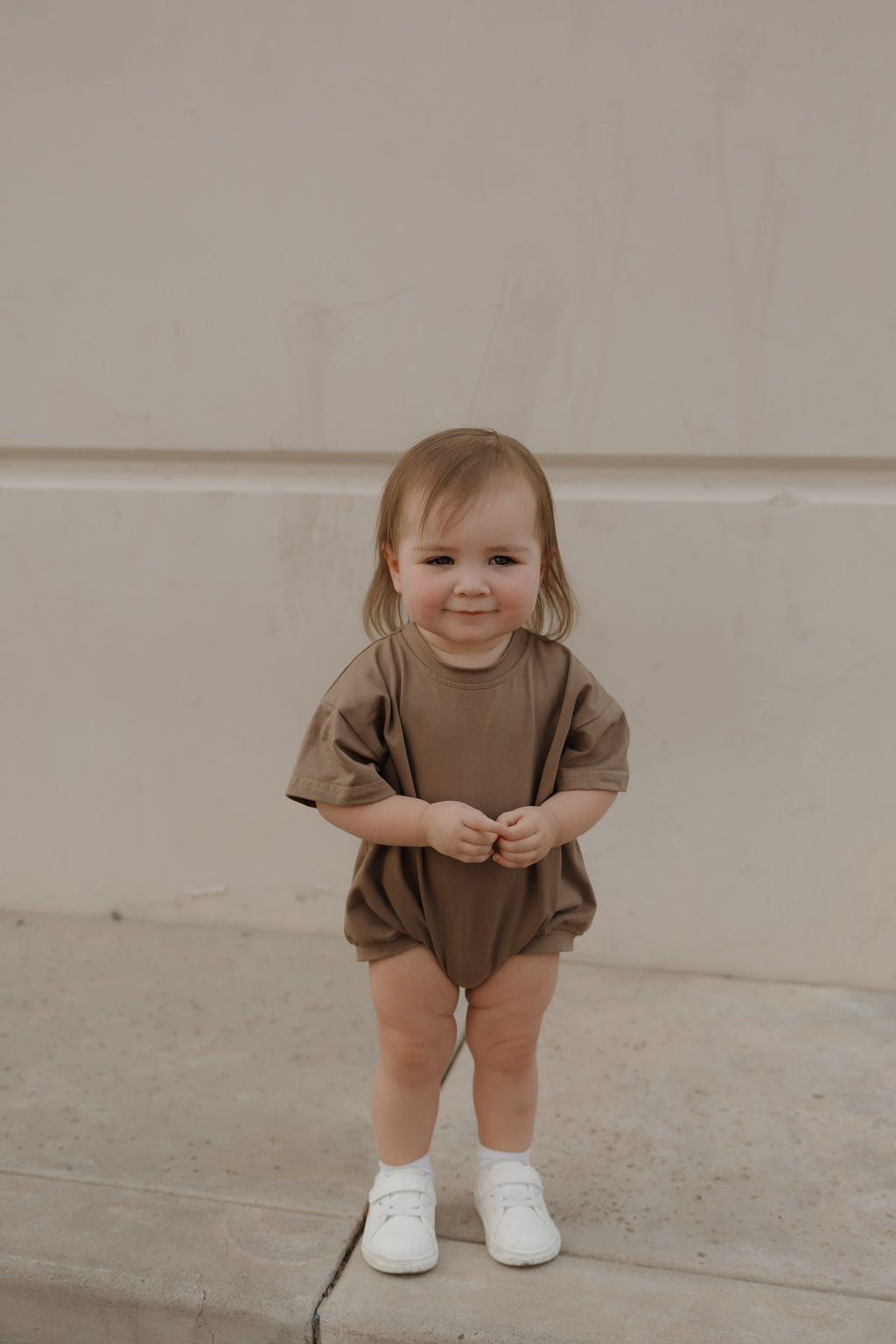 A toddler with light brown hair smiles at the camera, wearing the forever french baby Short Sleeve Romper in Patina and white shoes, standing on concrete against a beige wall.