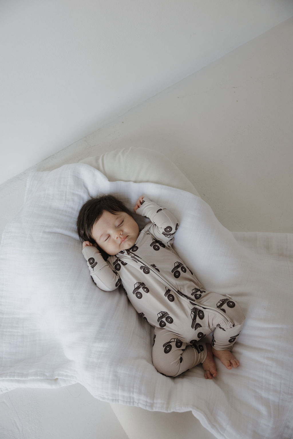 A baby sleeps peacefully on a white blanket, wearing forever french baby Bamboo Zip Pajamas in the Monster Truck print. The breathable fabric keeps them comfy, with their arms above their head and dark hair softly resting.