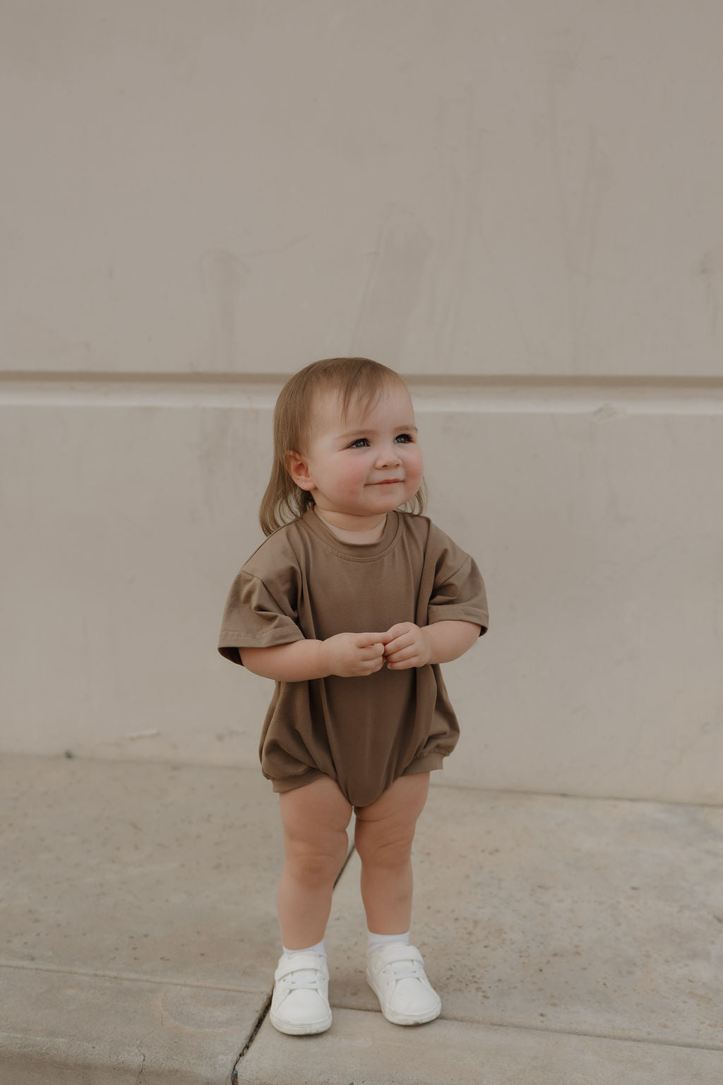 A young toddler with light brown hair, wearing the forever french baby Short Sleeve Romper | Patina and white shoes, stands on a concrete surface in front of a beige wall, looking up with a slight smile.