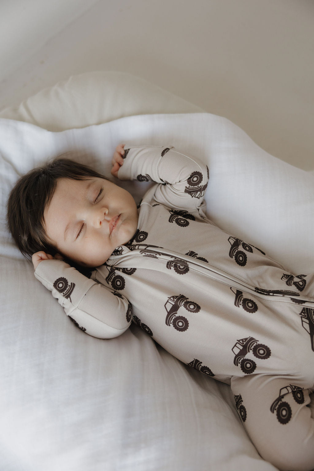 A baby with dark hair sleeps peacefully on a white pillow, dressed in forever french baby's Bamboo Zip Pajamas | Monster Truck—a beige, breathable, hypo-allergenic onesie patterned with black trucks.