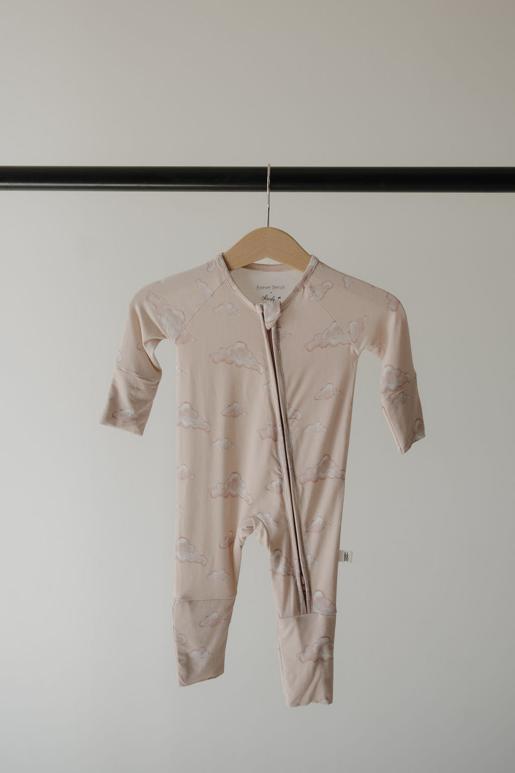 The Kendy 3 Bamboo Zip Pajamas | Ruby, in pale pink with a cloud pattern, hang on a wooden hanger. Made from breathable fabric, these long-sleeve, footed baby rompers feature a diagonal zipper from neck to leg.