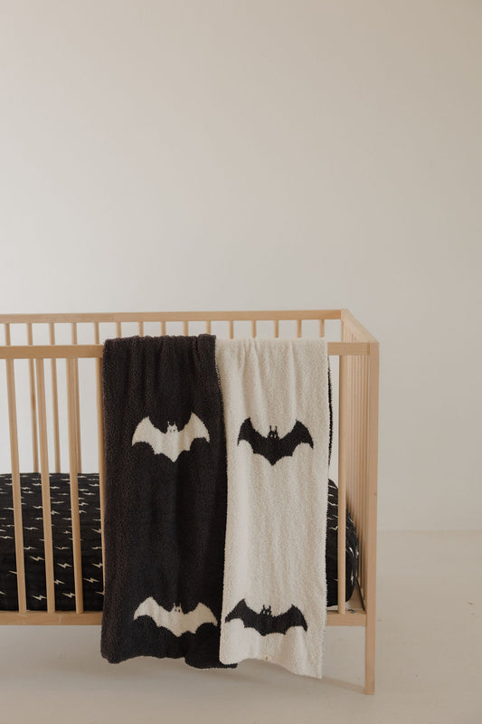 A minimalist wooden crib with a black mattress features two forever french baby Plush Blanket | It's Bats!—one in black and one in white—draped over the side, both OEKO-TEX certified and adorned with bat designs. The background is a plain off-white wall.