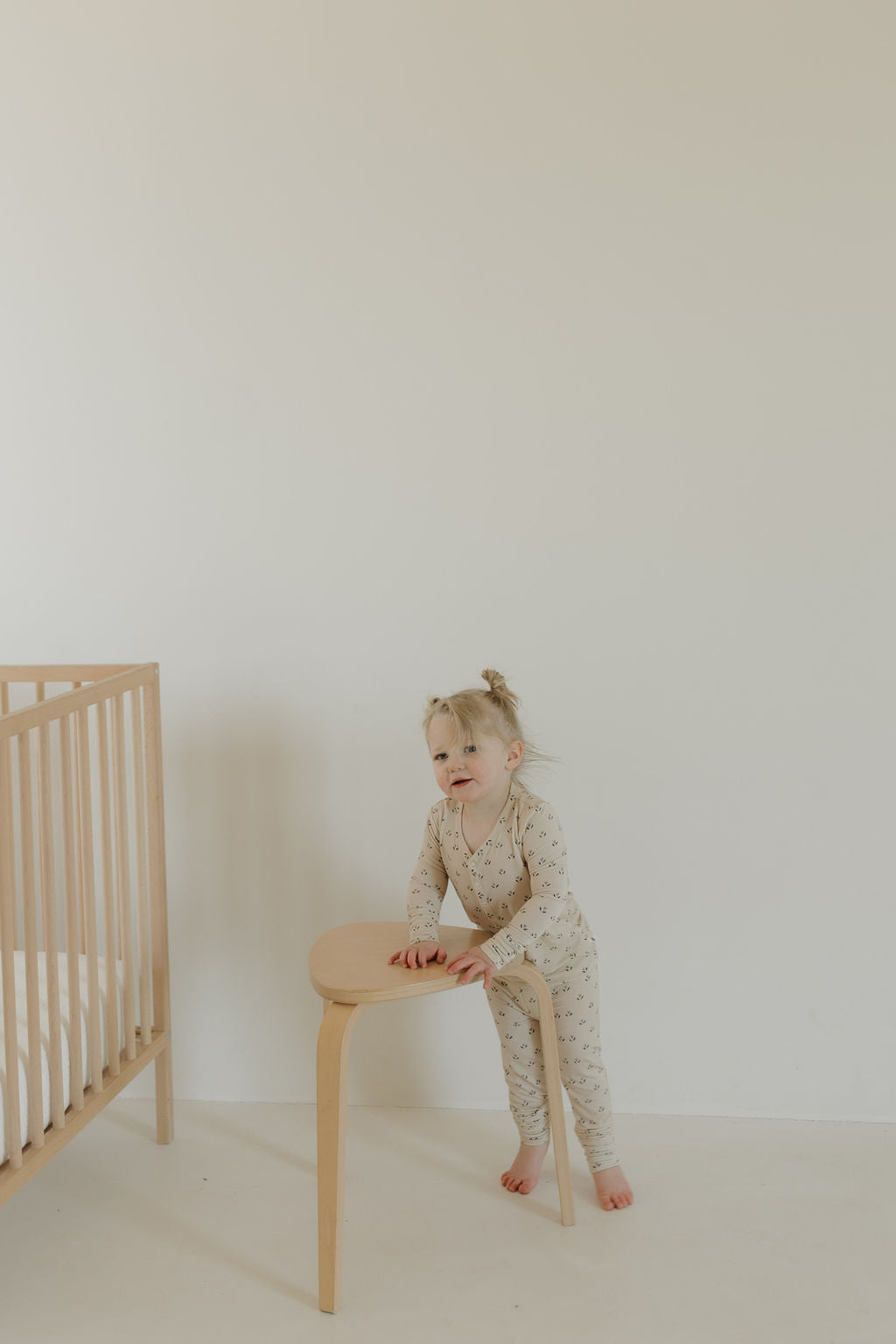 A toddler wearing the Bamboo Two Piece Pajamas in Flora by forever french baby stands next to a light wooden chair with a crib on the left. A plain white wall in the background creates a minimalist and cozy atmosphere.