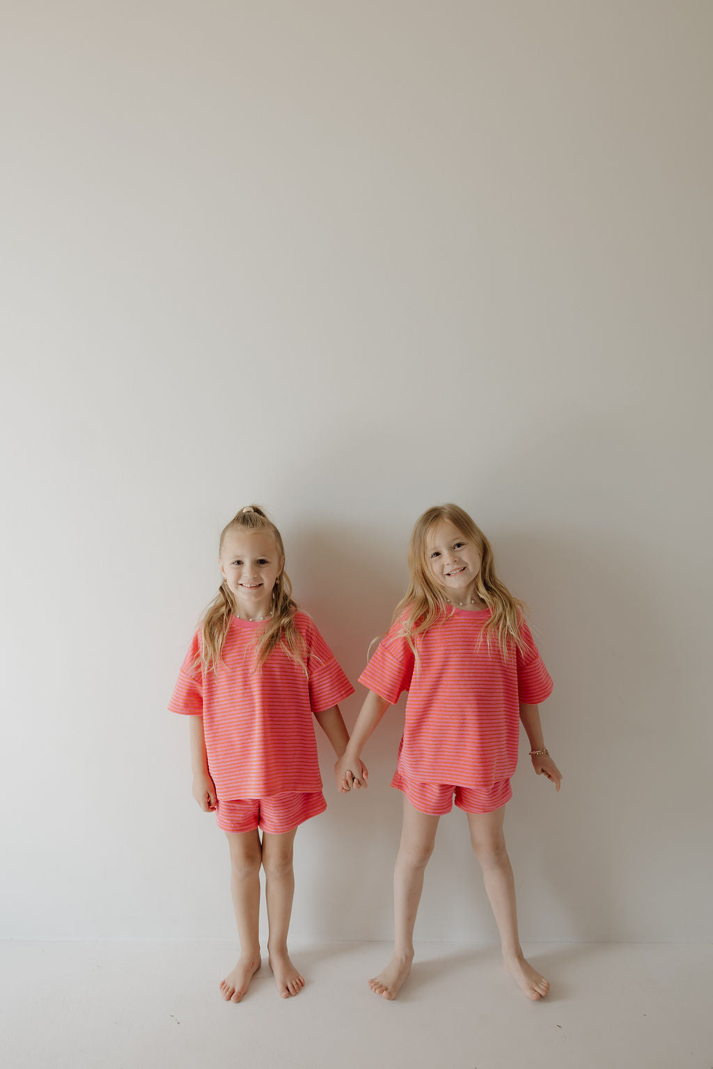 Two young girls stand barefoot against a white wall, holding hands and smiling in matching forever french baby Child Terry Short Set | Strawberry outfits—highlighting the joy of family in coordinated style.
