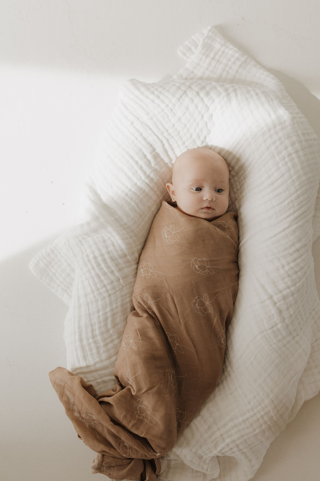 A baby lies with a calm expression on a soft, white quilted surface, wrapped in the forever french baby Muslin Swaddle in Sweet Dreams Floral. The 100% muslin cotton blanket offers extra comfort as soft, diffused light surrounds them.