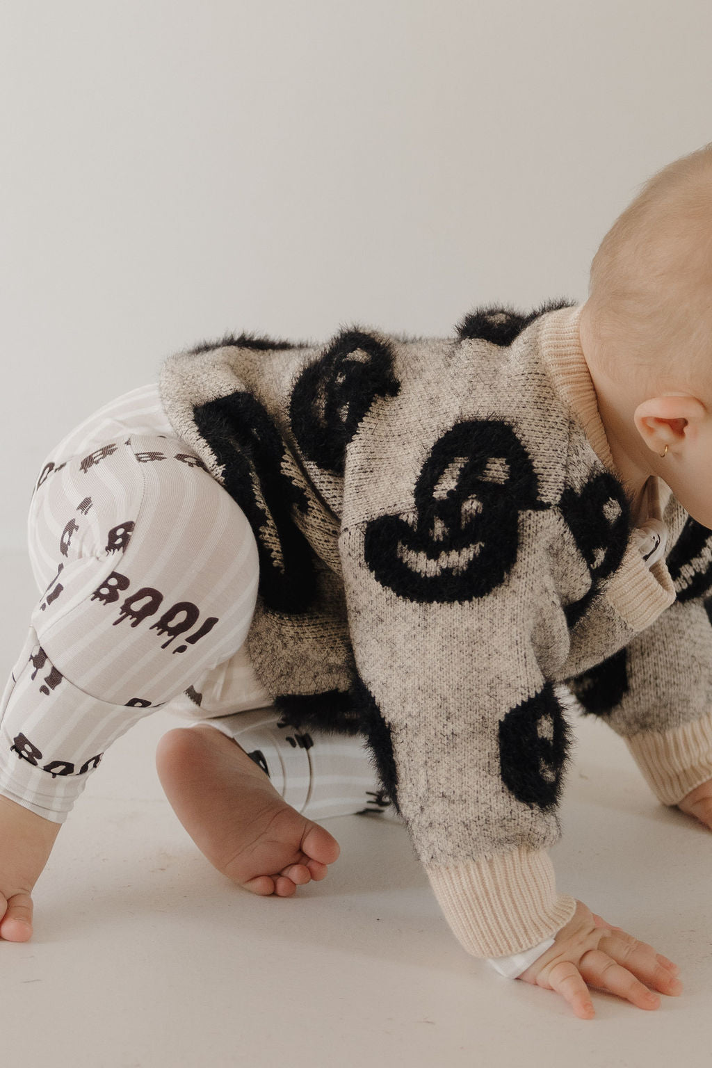 A baby from forever french baby crawls on a light floor, wearing the Cozy Sweater | Halloween—a fuzzy beige piece with bold black smiley faces—plus white "BOO!" leggings; their face is out of view.