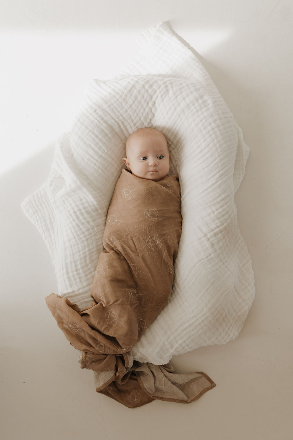 A baby is wrapped in a forever french baby Muslin Swaddle | Sweet Dreams Floral, lying on white textured fabric. The 100% muslin cotton wrap gently cradles the infant, gazing upwards. Soft lighting casts shadows around the baby, highlighting serene innocence.