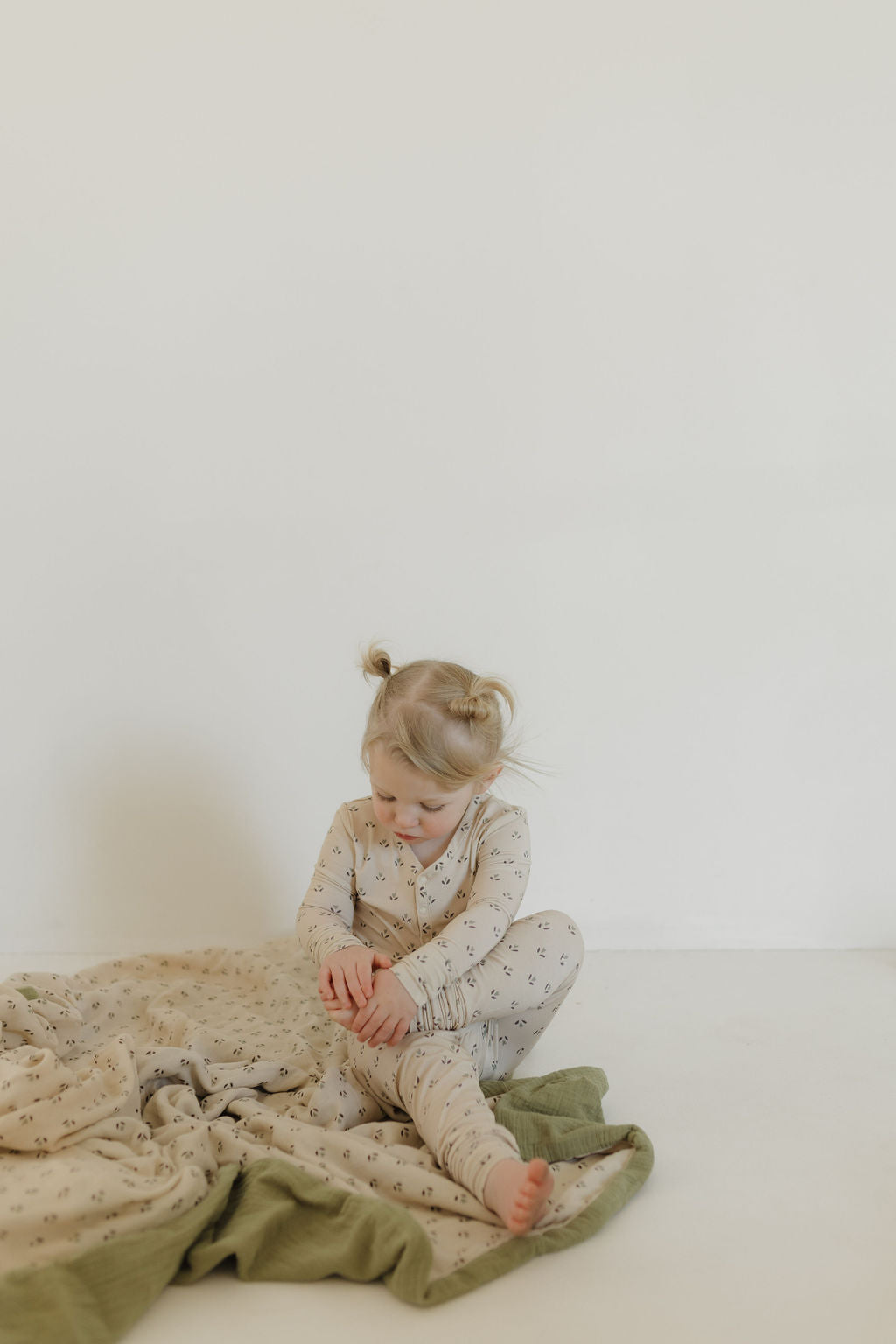 A young child with light hair sits on the floor wearing forever french baby's Bamboo Two Piece Pajamas in Flora, featuring small patterns. They are surrounded by a soft, light and dark green blanket against a plain background.