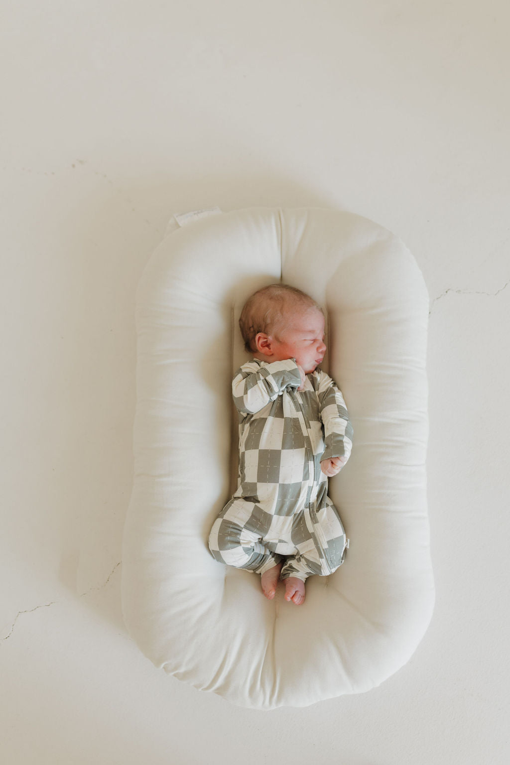 A newborn sleeps peacefully on a soft, white cushion, wrapped in hypoallergenic clothing. The little one wears "Bamboo Zip Pajamas | Oat & Sage" by forever french baby, comfortably resting on a light-colored surface.