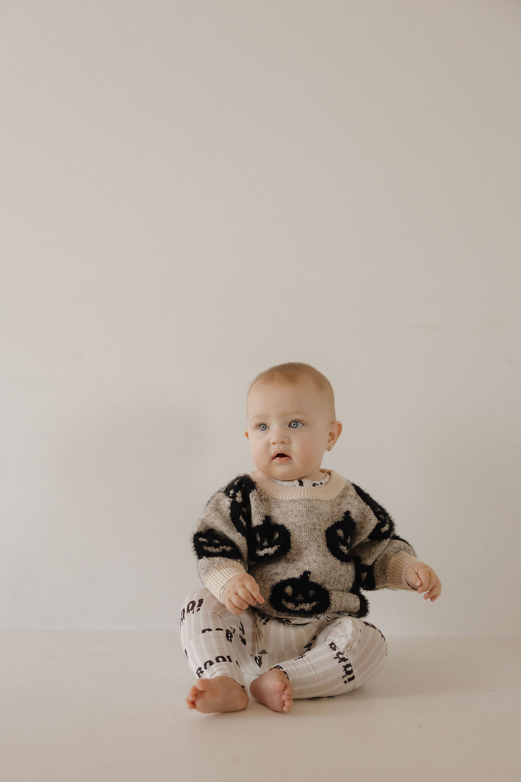 A baby leans against a light wall in the forever french baby Cozy Sweater | Halloween—beige with black smiley faces—for a cozy, festive look.