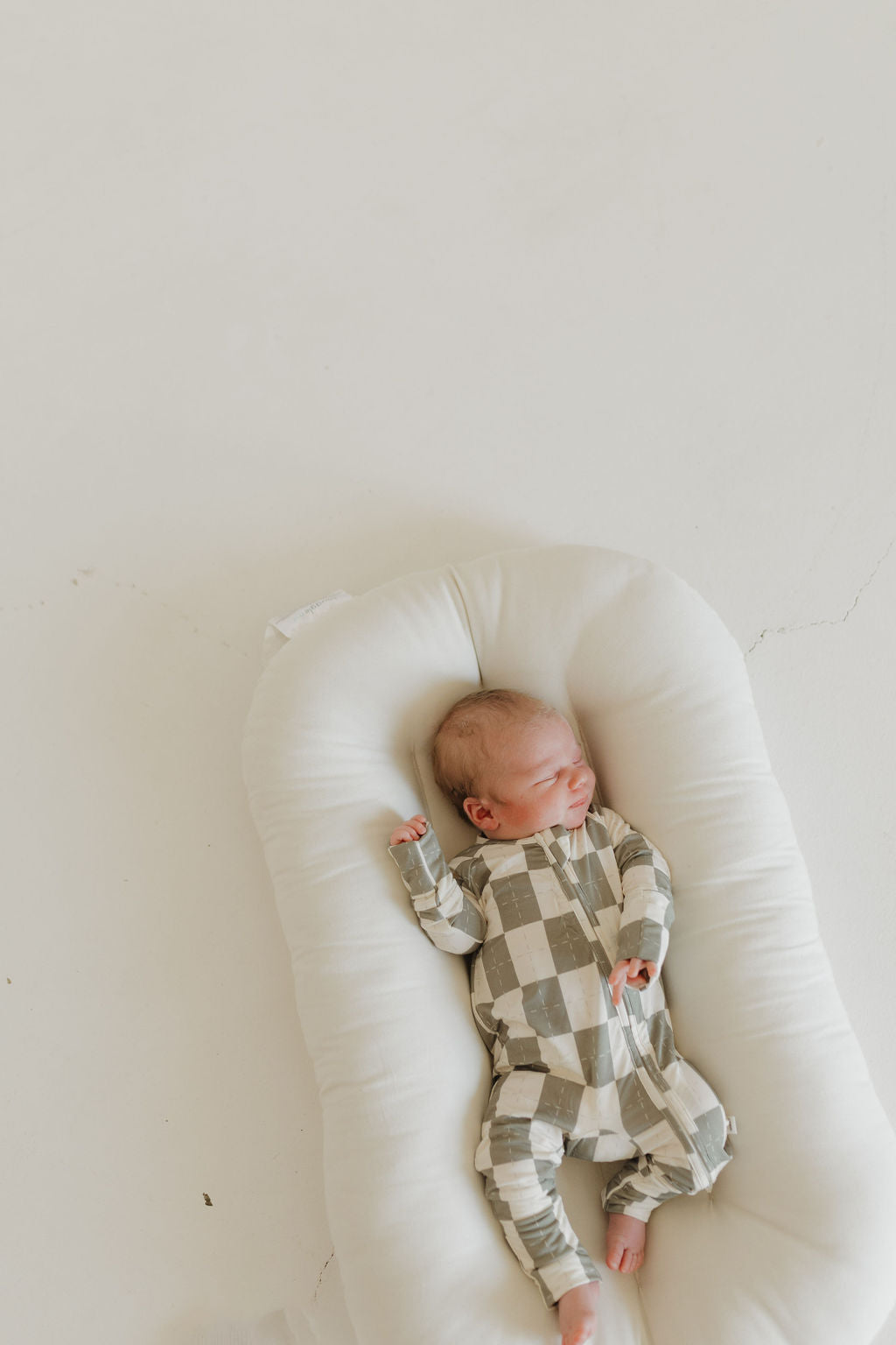 A baby, serene in forever french baby's Oat & Sage Bamboo Zip Pajamas, sleeps on a soft white cushion against a light backdrop. The gentle colors of the hypoallergenic pajama contrast softly with the floor as the baby's arms lay slightly raised.