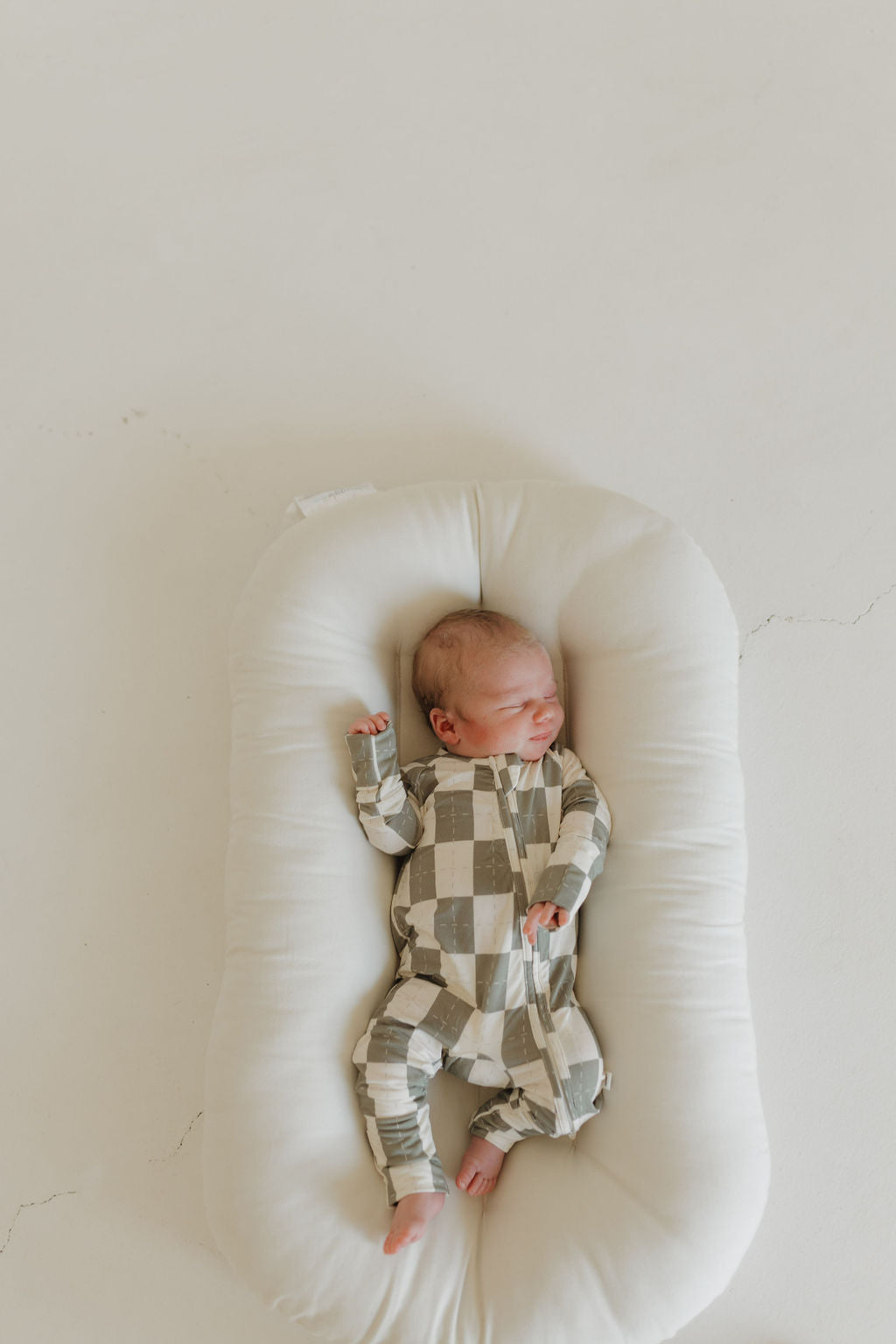 A baby relaxes on a soft white cushion in forever french baby's Bamboo Zip Pajamas in Oat & Sage. The cozy, breathable, hypo-allergenic pajamas feature a green and white checkered pattern, keeping the baby peaceful against the light background.