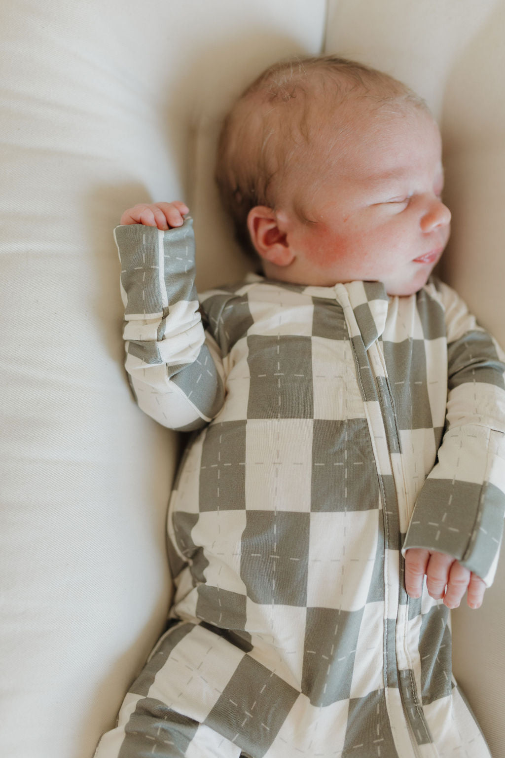 A baby sleeps peacefully in forever french baby's Bamboo Zip Pajamas | Oat & Sage on a soft, light-colored surface. The green and white checkered onesie is crafted from hypo-allergenic bamboo, with the baby lying on their back and one hand beside their head.