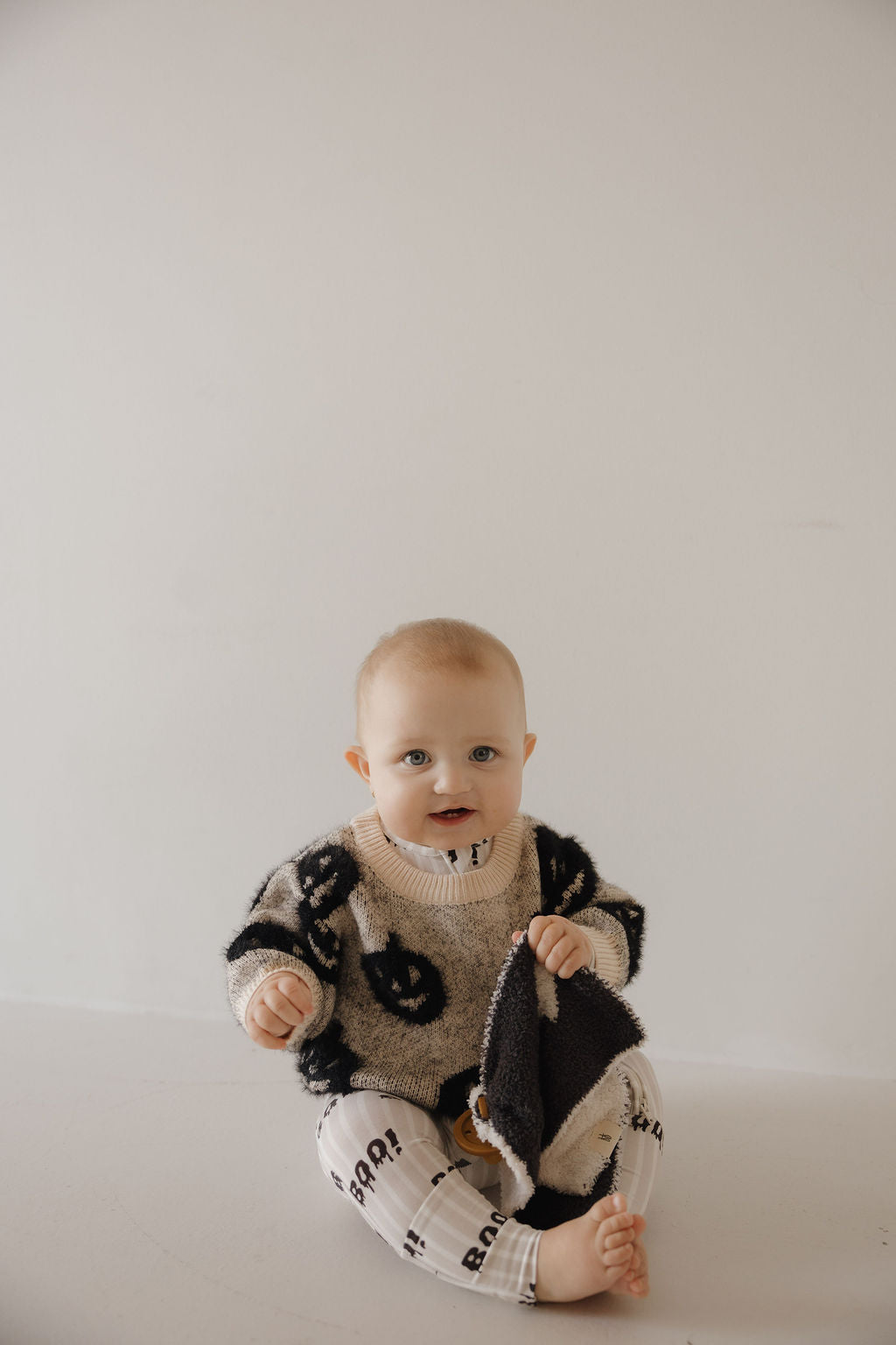 A baby sits barefoot on a white floor, wearing the forever french baby Cozy Sweater | Halloween and patterned "BOO!" pants, holding a soft black and white blanket while looking at the camera.