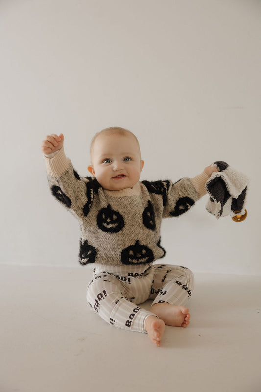 A baby sits on the floor against a plain background, smiling with arms raised. Dressed in the forever french baby Cozy Sweater | Halloween, the baby holds a small black-and-white blanket and wears white pants with lettering.
