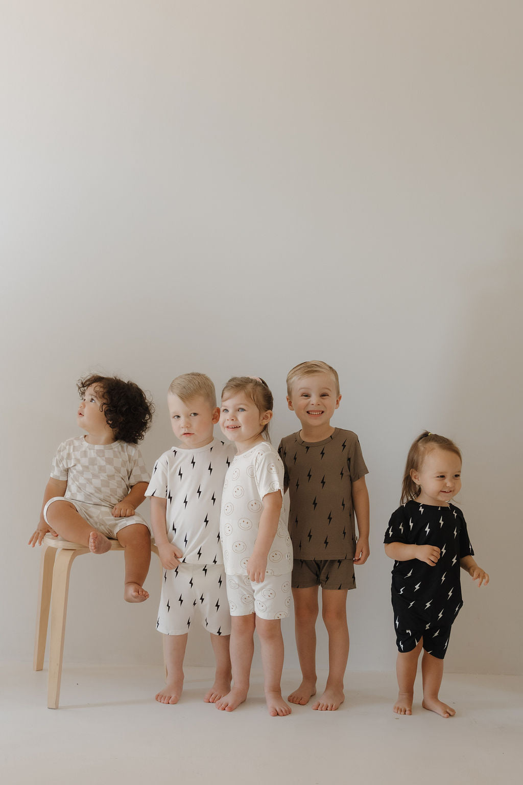 Five young children pose against a light background, wearing forever french baby's Bamboo Short Set in Brown & Black Lightning Bolt—neutral, hypo-allergenic outfits with simple patterns. One child sits on a wooden stool while the others stand barefoot.