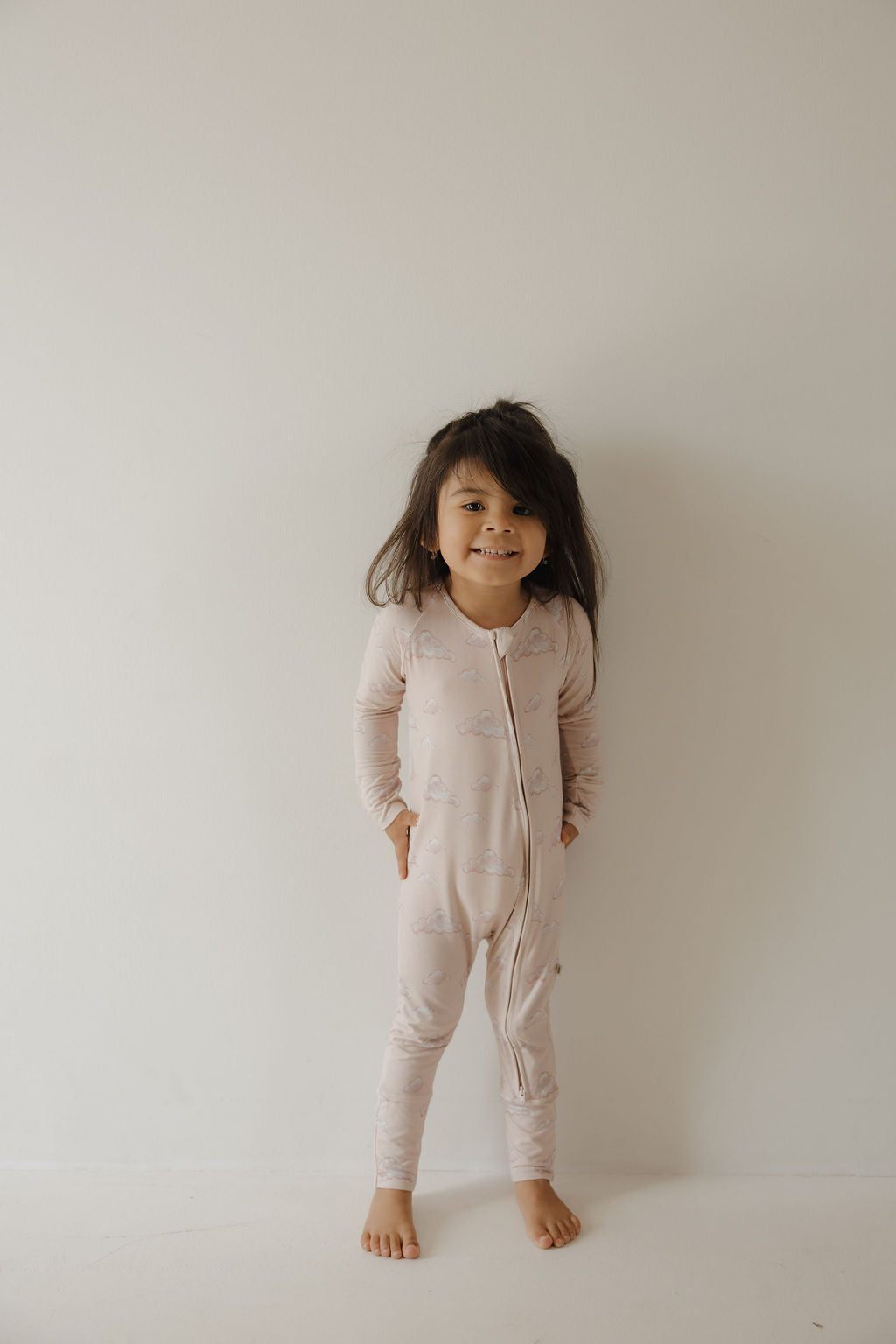 A young child with long dark hair stands barefoot against a plain wall, smiling and wearing Kendy 3’s Bamboo Zip Pajamas | Ruby—a light pink, long-sleeved, zip-up bamboo onesie with a subtle cloud pattern.