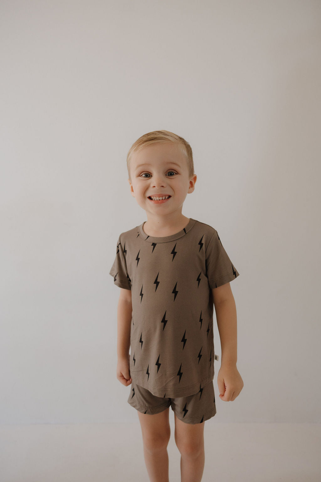 A young child with short blonde hair smiles against a plain background, wearing the forever french baby Bamboo Short Set in brown with a black lightning bolt pattern—stylish, hypo-allergenic comfort for kids.