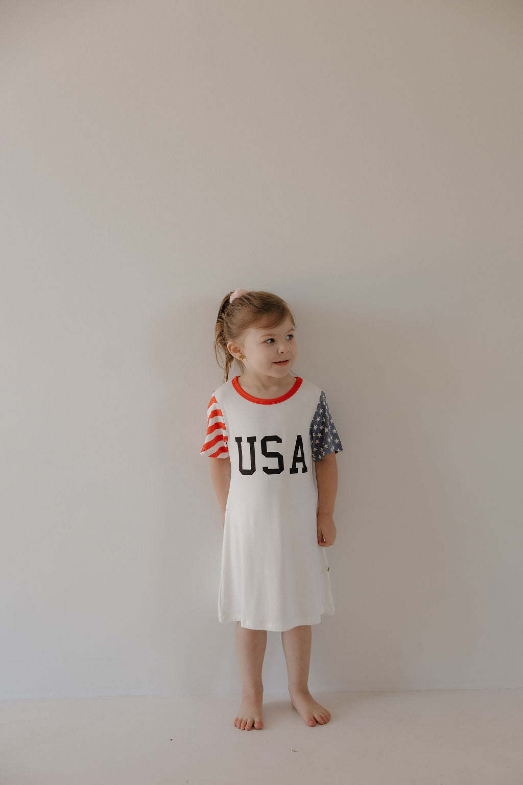 A young girl stands barefoot against a plain wall, wearing the forever french baby Bamboo T-Shirt Dress | Stars & Stripes, featuring "USA" on the front with one red and white striped sleeve and one blue sleeve adorned with white stars.