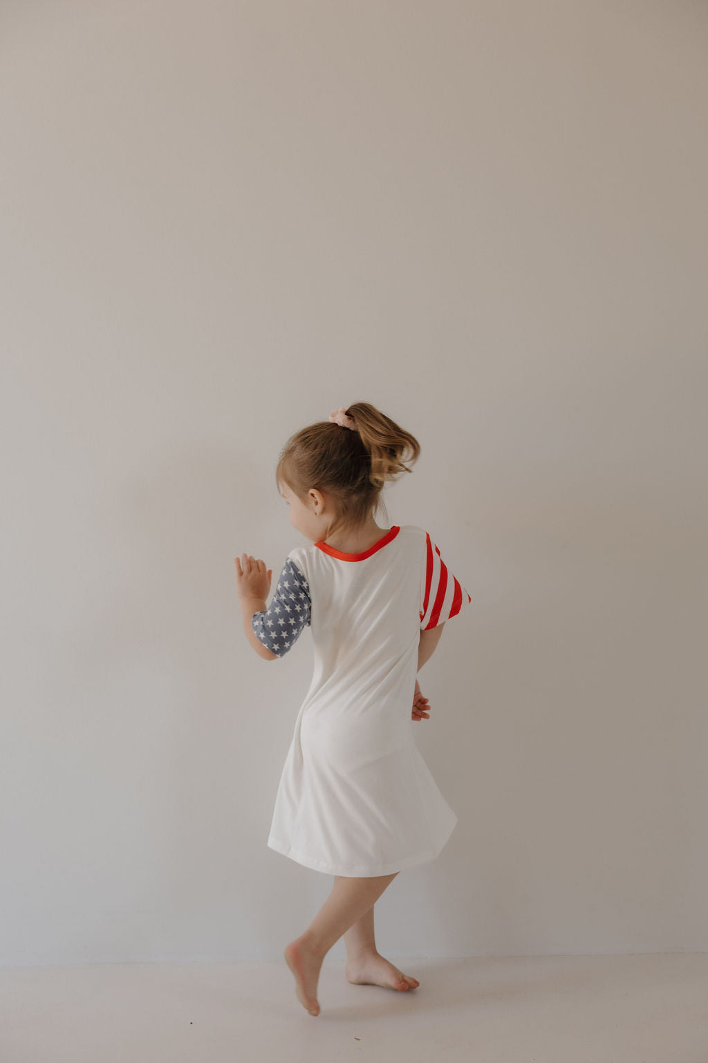 A young girl with a ponytail wears the forever french baby Bamboo T-Shirt Dress | Stars & Stripes, featuring an American flag-inspired design. She stands barefoot against a plain light wall, turned slightly away from the camera.