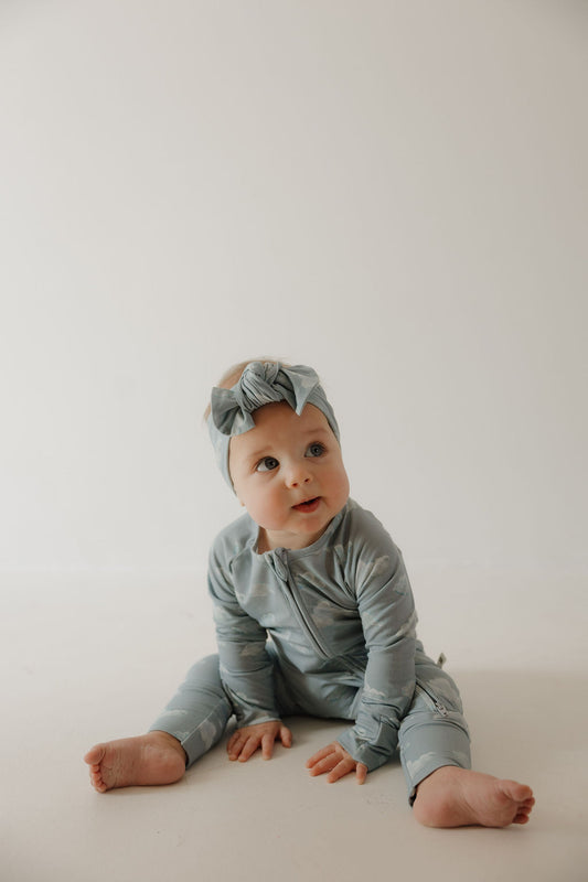 A baby in Kendy 3’s Bamboo Zip Pajamas | Duke, with a matching headband, sits on the floor against a light backdrop, looking up curiously. The soft bamboo fabric offers breathable comfort for sensitive skin.