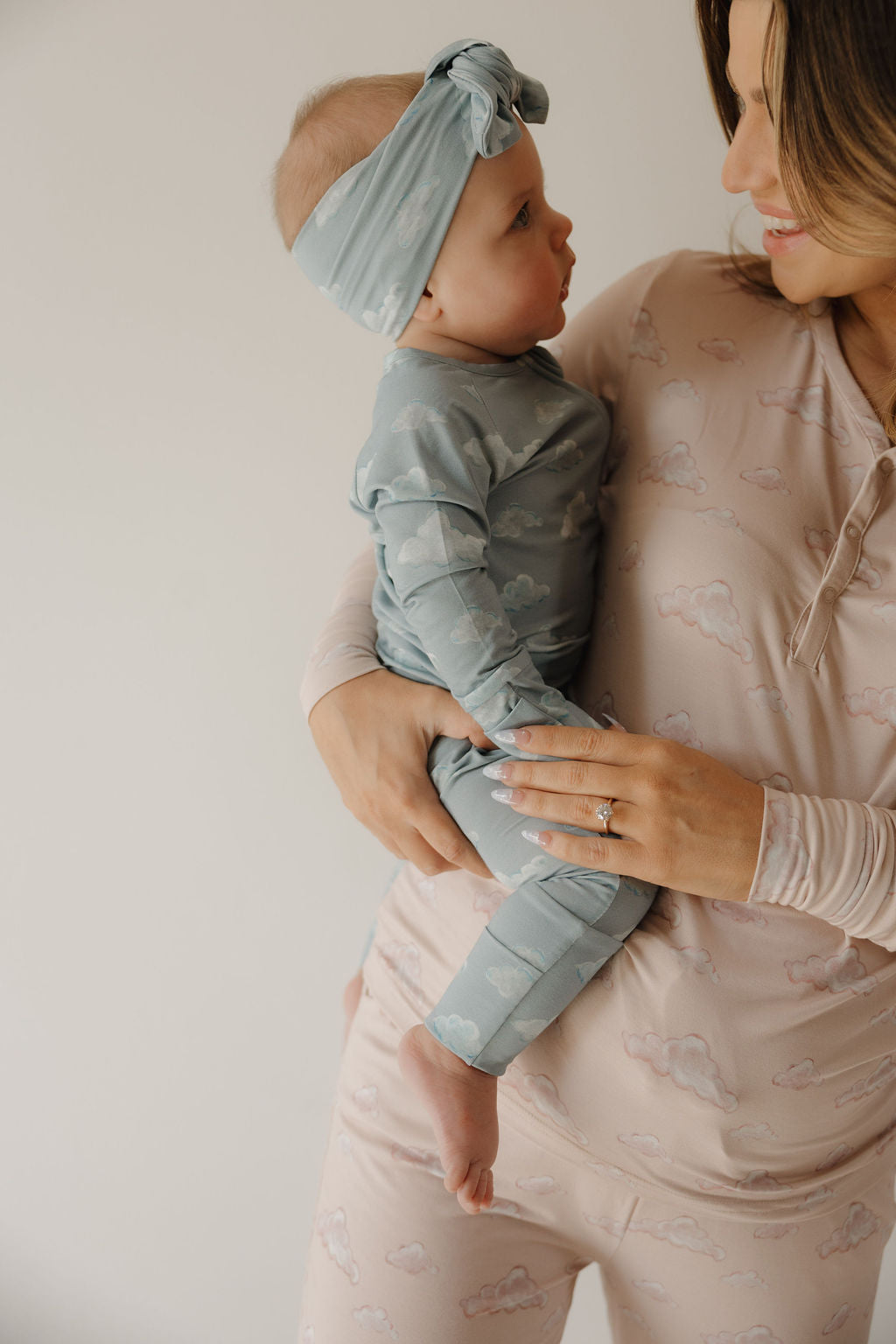 A woman in a light pink cloud-patterned outfit smiles at a baby wearing matching blue clouds and a Bamboo Head Wrap | ff x kendy by Kendy 3, both showcasing their adorable style against a plain background.