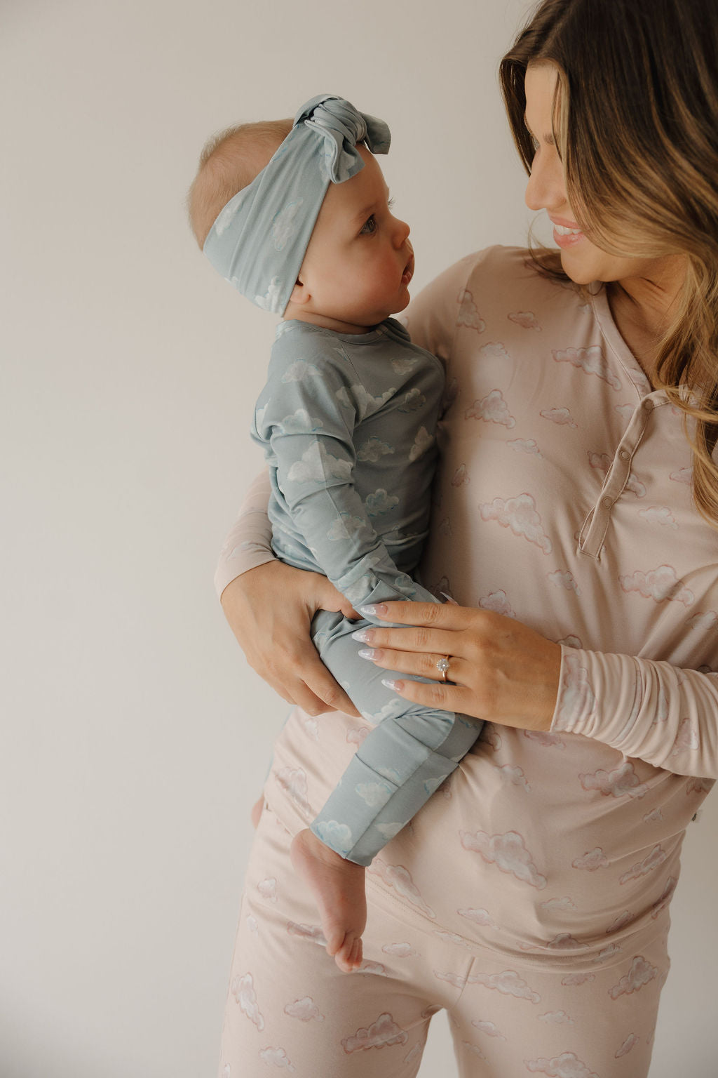A woman in cloud pajamas holds a baby in green pajamas with a Kendy 3 Bamboo Head Wraps | ff x kendy. They smile at each other.