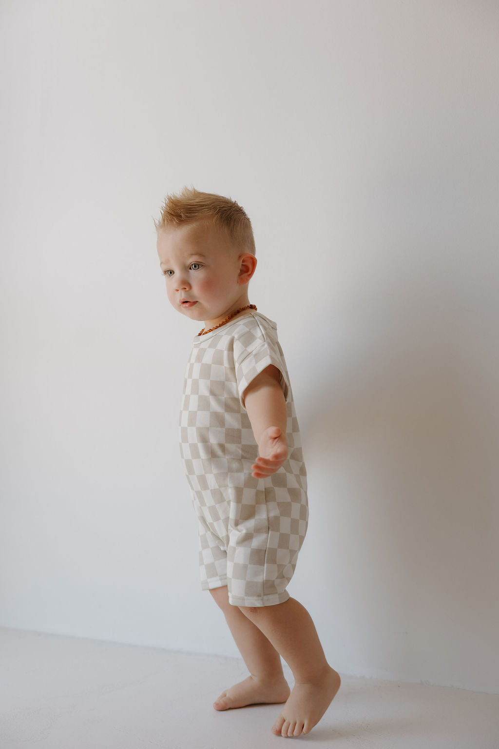 A young child (newborn–24 months) stands barefoot by a white wall, wearing the forever french baby French Terry Romper | Checkerboard, a light short-sleeve romper with beige and white checks and snap closures.