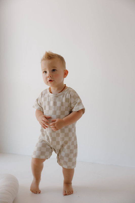 A toddler with short blonde hair stands barefoot on a white floor, wearing the forever french baby French Terry Romper | Checkerboard— a breathable beige and white checkered outfit with snap closures and a beaded necklace, set against a plain white background.