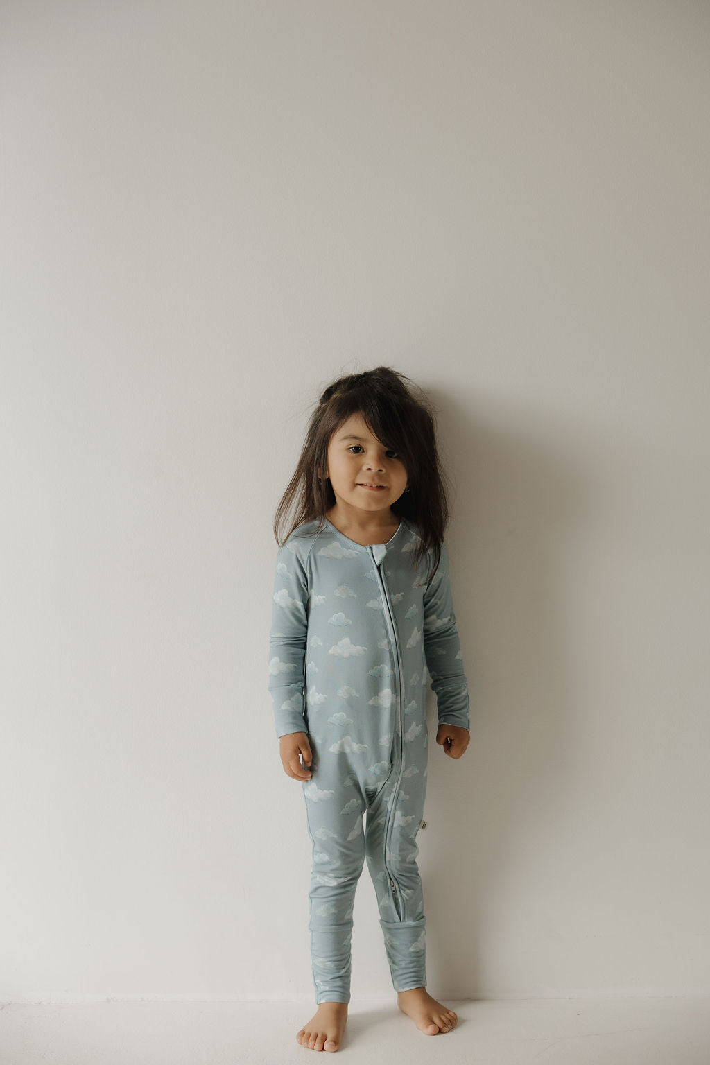 A young child with long dark hair stands barefoot against a white wall, wearing Kendy 3's Bamboo Zip Pajamas | Duke—light blue bamboo pajamas with a cloud pattern and front zipper, designed for breathable, all-night comfort.