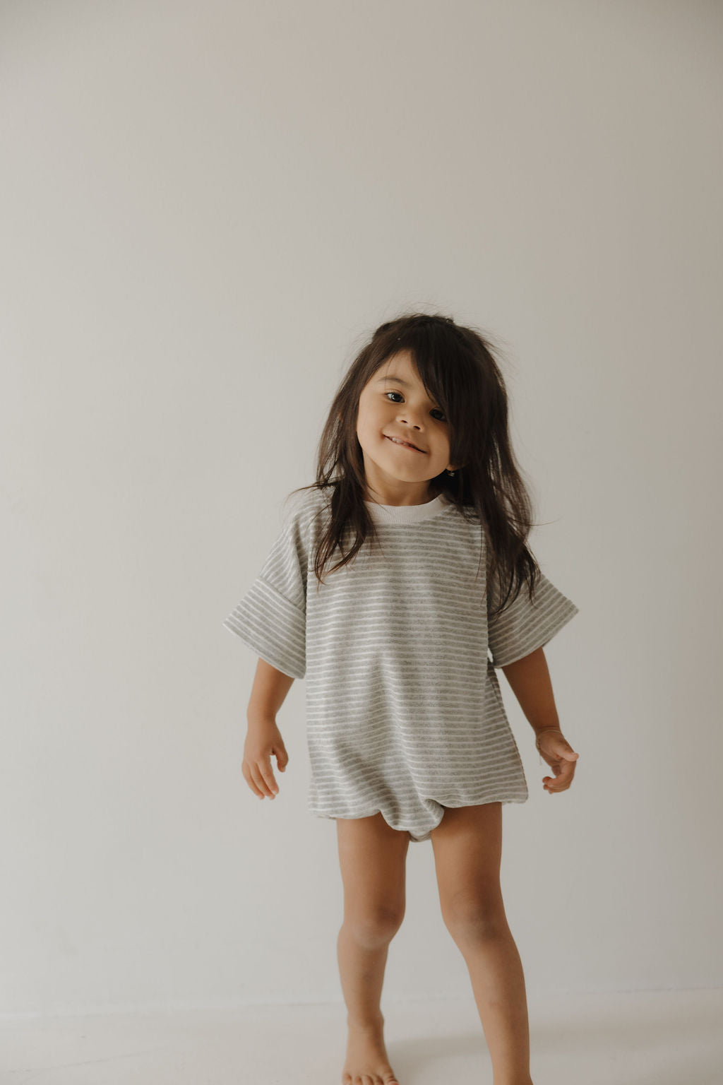 A young child with long dark hair smiles slightly, barefoot against a plain background, wearing the forever french baby Terry T-Shirt Romper in Grey Stripe—perfect for easy diaper changes and special family moments.