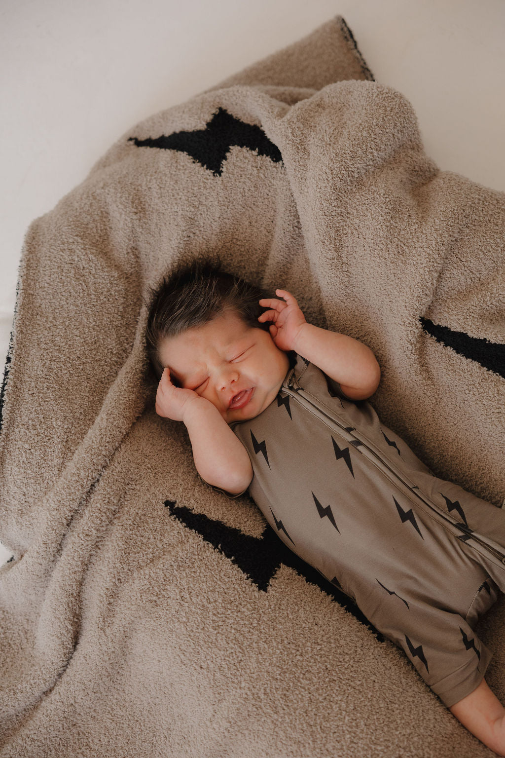 A newborn in forever french baby's Bamboo Zip Shortie | Brown & Black Lightning Bolt sleeps peacefully on a tan blanket with coordinating lightning bolt designs, eyes closed and hands by the face.