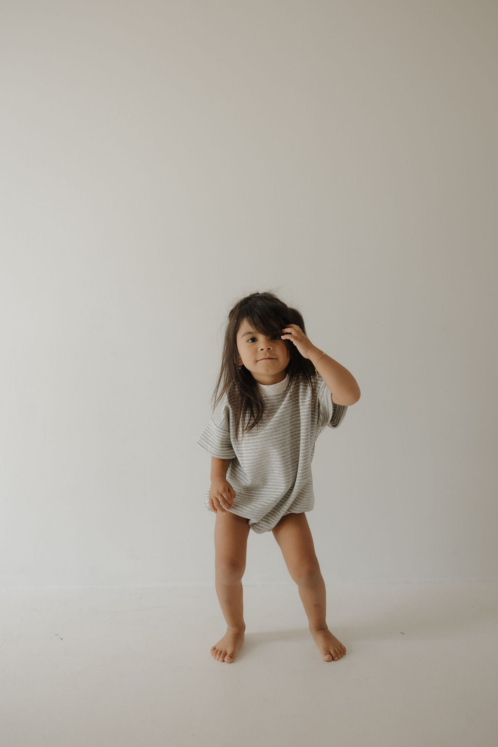 A young child with long dark hair stands barefoot against a plain light wall, wearing the forever french baby Terry T-Shirt Romper in Grey Stripe, looking at the camera and touching their head—capturing sweet family moments.