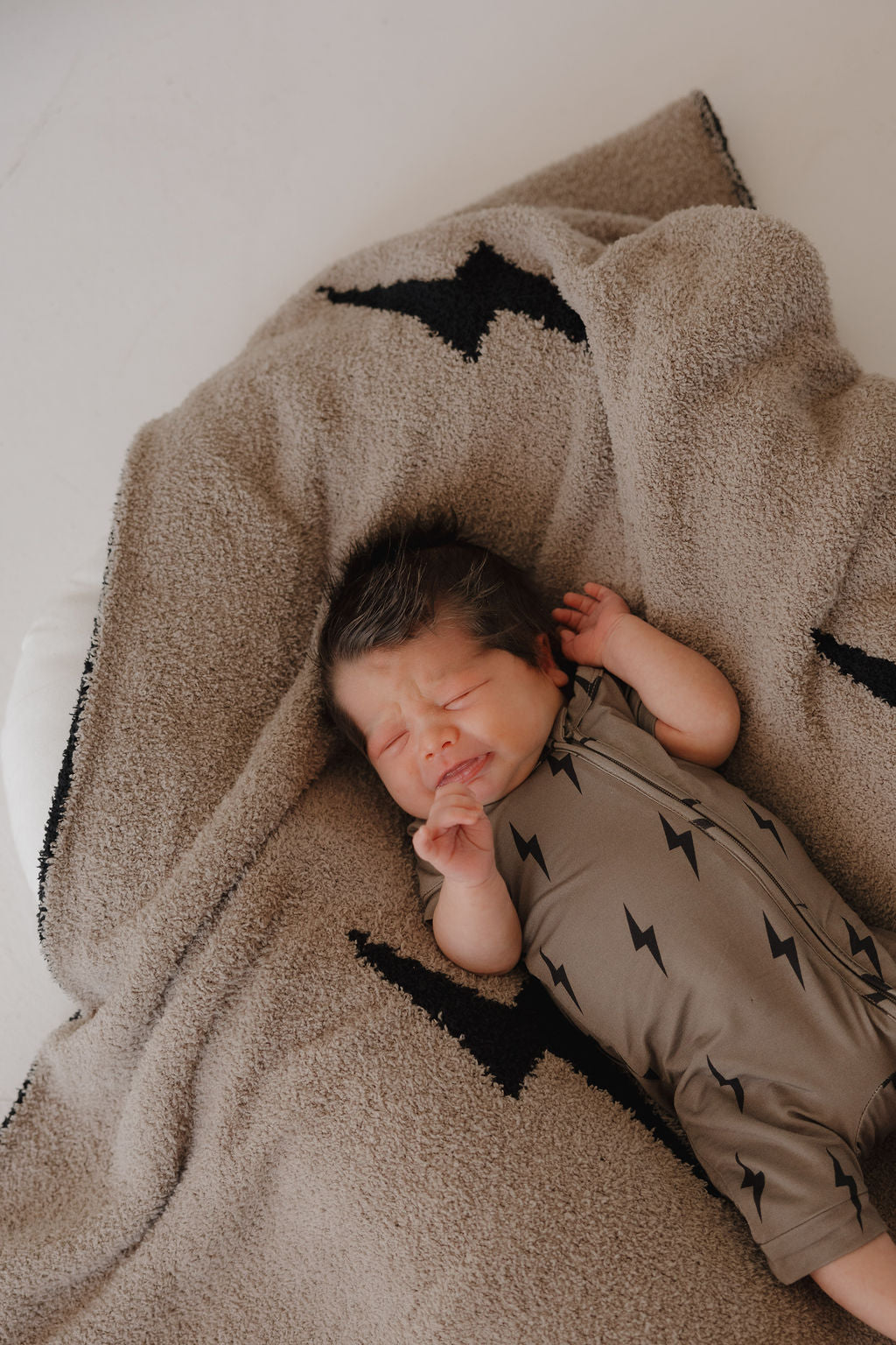 A baby with dark hair sleeps on a beige blanket with black lightning bolts, wearing forever french baby's Bamboo Zip Shortie in Brown & Black Lightning Bolt for safe, gentle comfort.