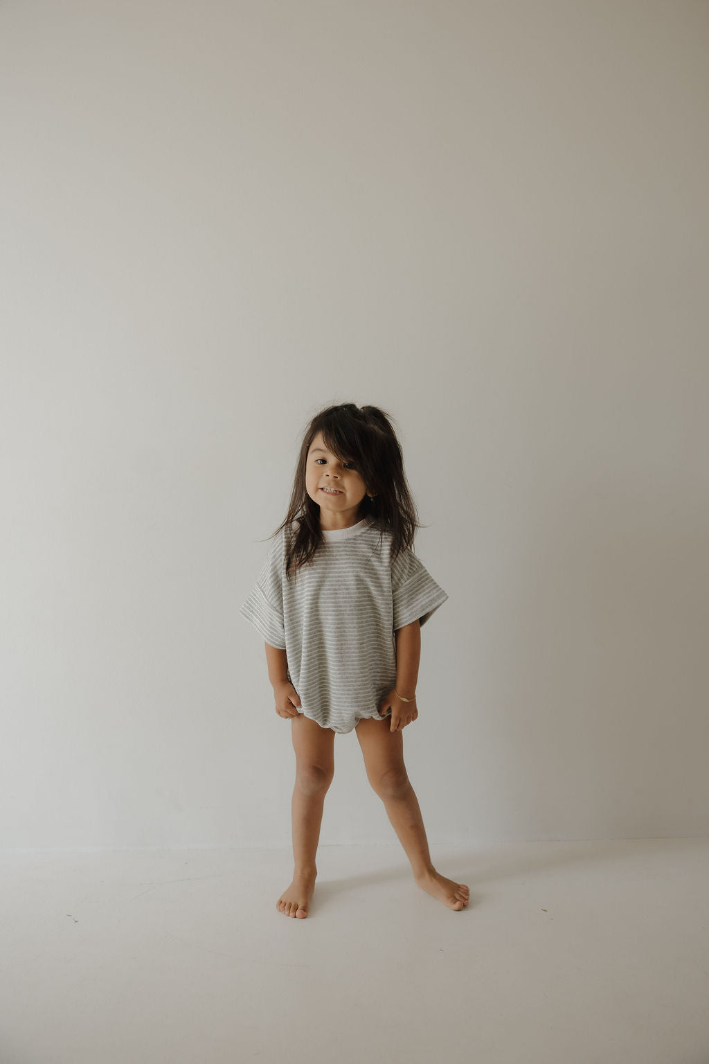 A young child with long dark hair stands barefoot against a beige wall, wearing the forever french baby Terry T-Shirt Romper in Grey Stripe. The child gazes off to the side, capturing a relaxed, everyday family moment.