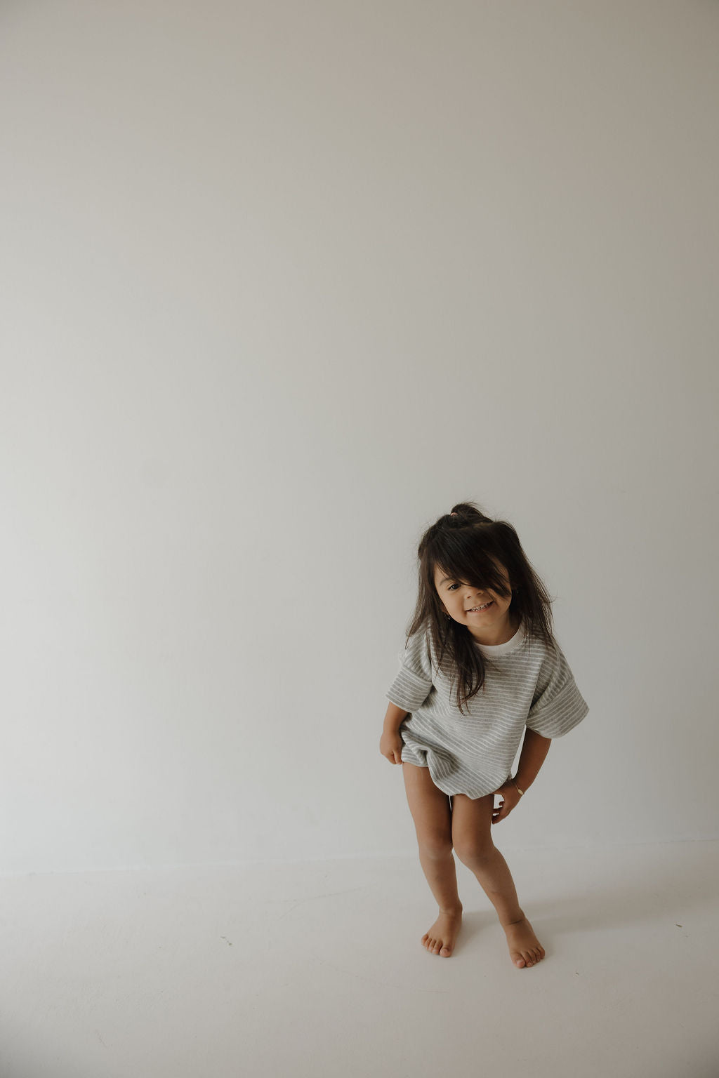 A young child with long dark hair wears the forever french baby Terry T-Shirt Romper in Grey Stripe, standing barefoot against a plain white wall and smiling playfully.