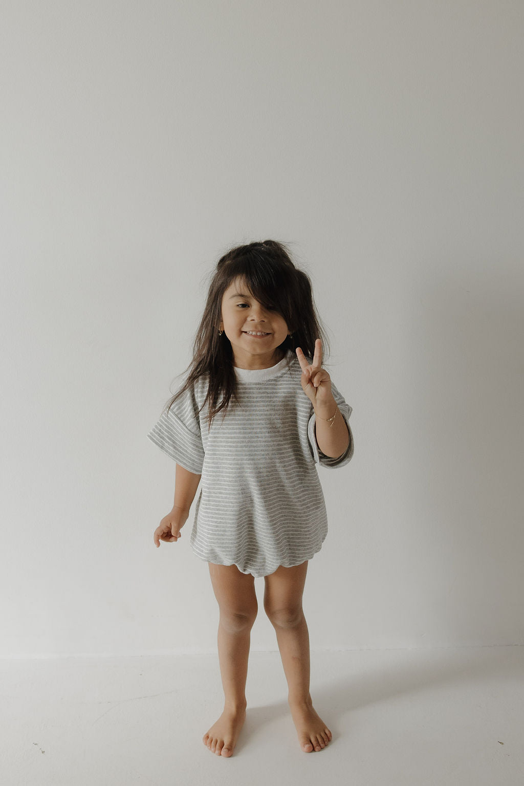 A young child with long dark hair stands barefoot against a white wall, cheerfully flashing a peace sign in the forever french baby Terry T-Shirt Romper | Grey Stripe—perfect for capturing joyful family moments.