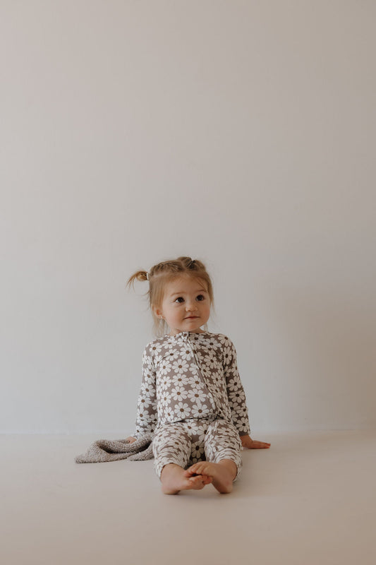 A young child with light hair in pigtails sits barefoot against a plain wall, wearing forever french baby Bamboo Zip Pajamas | Darlin', made from breathable bamboo fabric, and holding a small blanket.