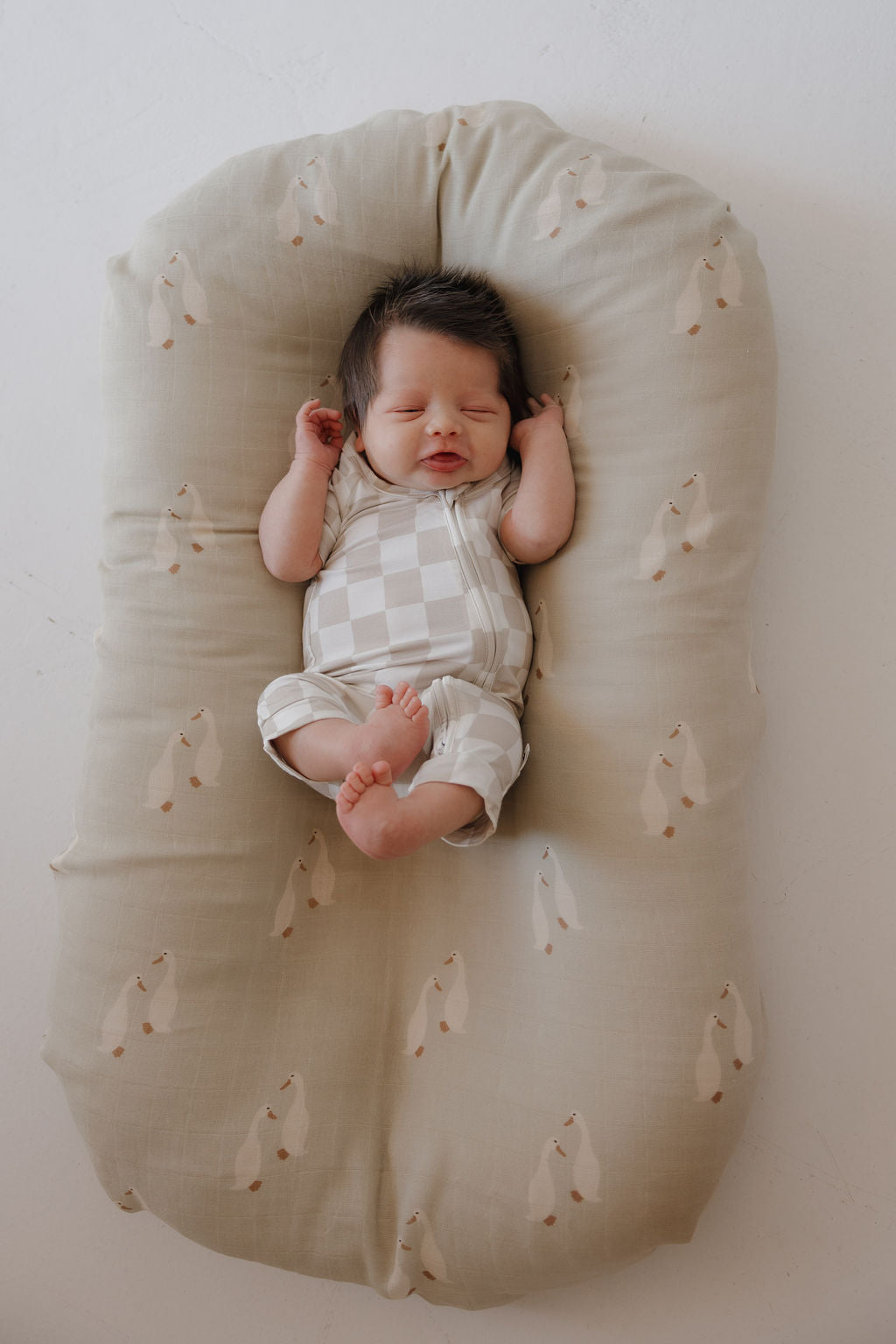A newborn with dark hair sleeps on a beige bear-print cushion, wearing the forever french baby Bamboo Zip Shortie | Checkerboard romper in breathable beige and white check, arms up and legs curled in comfort.