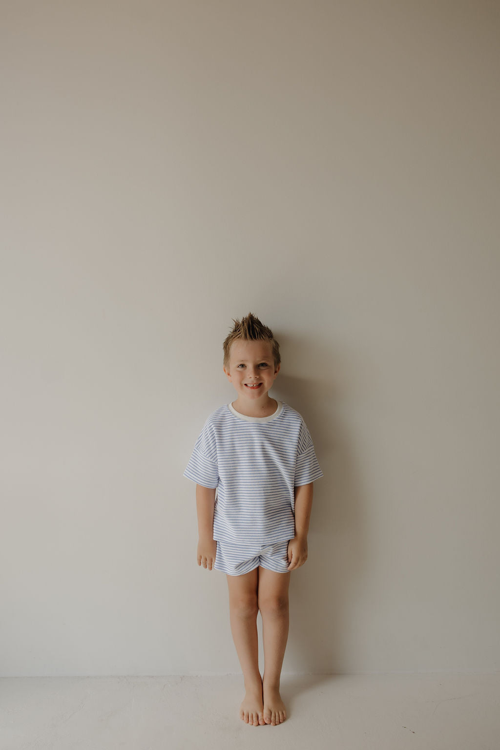 A young child with short, spiked blond hair stands barefoot against a plain beige wall, smiling in the forever french baby Child Terry Short Set | Ocean Stripe—perfect for capturing family moments.