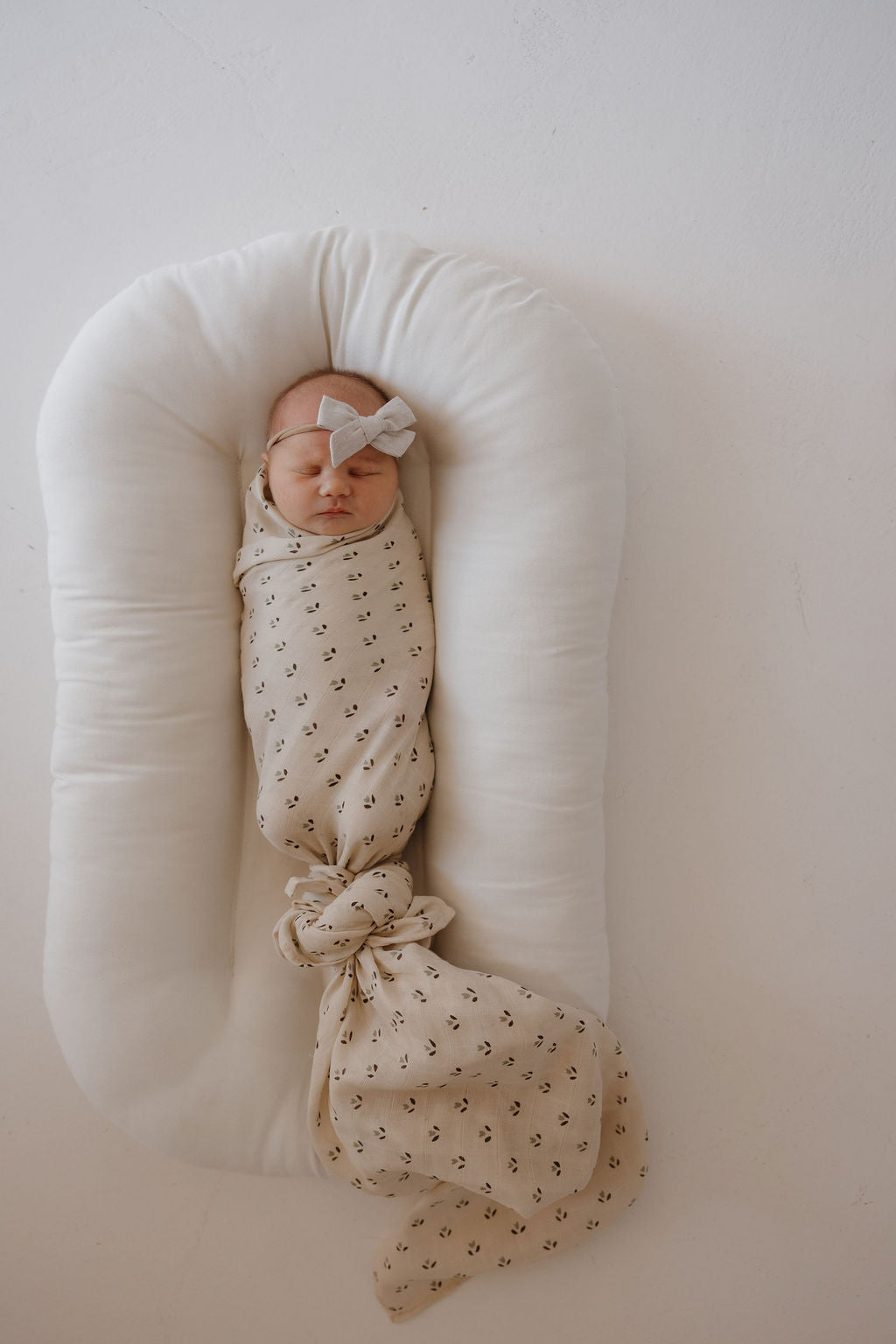 A newborn wrapped in the forever french baby Muslin Swaddle | Flora with a matching headband, lying on a white cushioned baby lounger against a plain background.